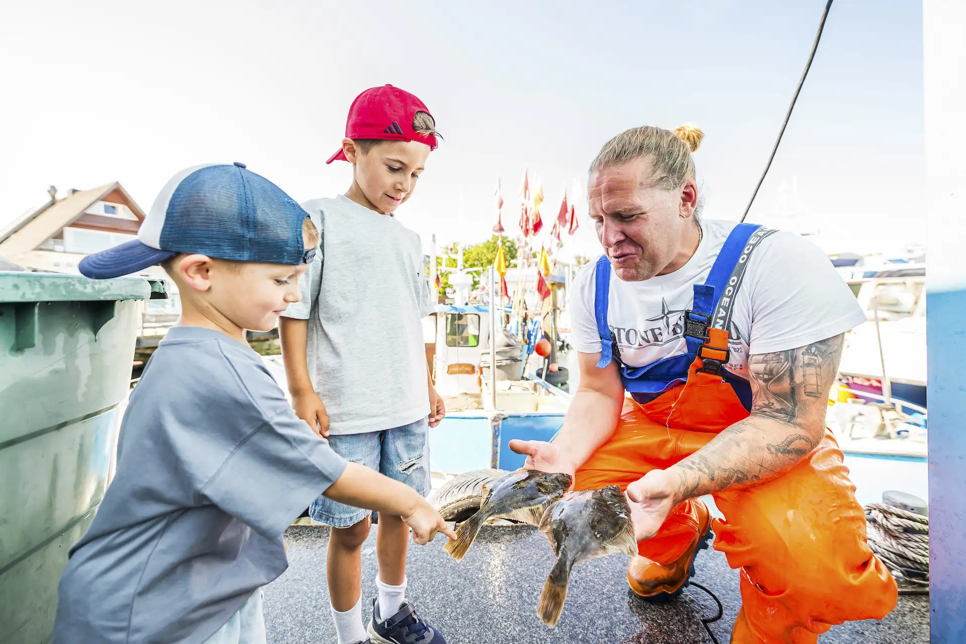 Ein Fischer zeigt zwei kleinen Jungs verschiedene Fische im Niendorfer Hafen.