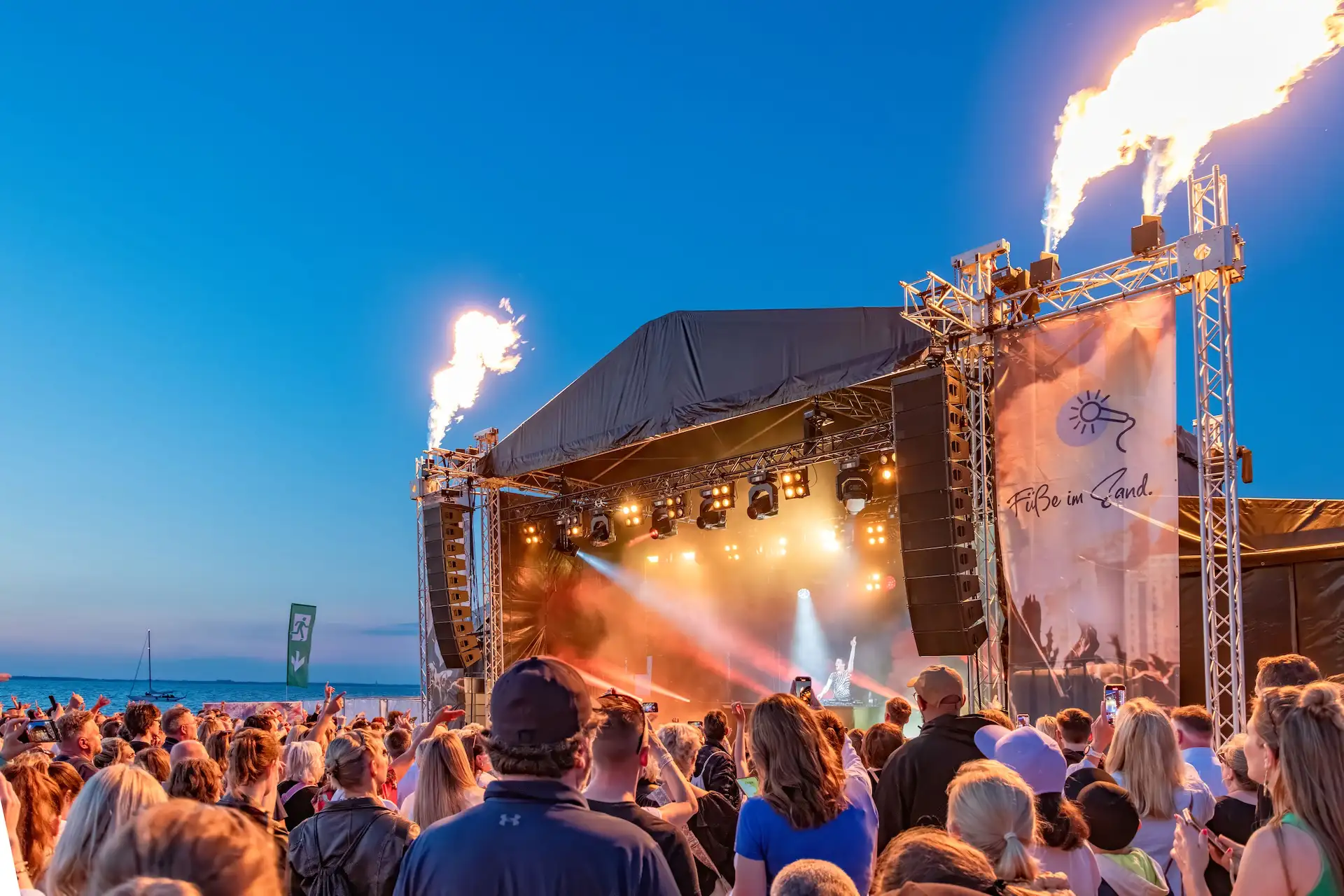 Eine große Menge Menschen steht vor einer Bühne, auf der eine Licht und Feuershow gezeigt wird, direkt am Strand.